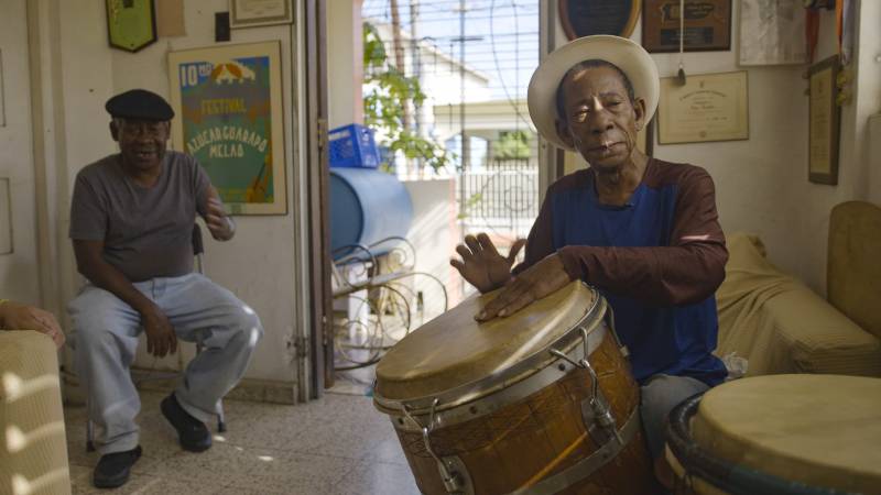 Men in an open-air room groove to the beat as Jesús Cepeda, who appears more prominently on the right side of the photo, plays a large barrel drum.