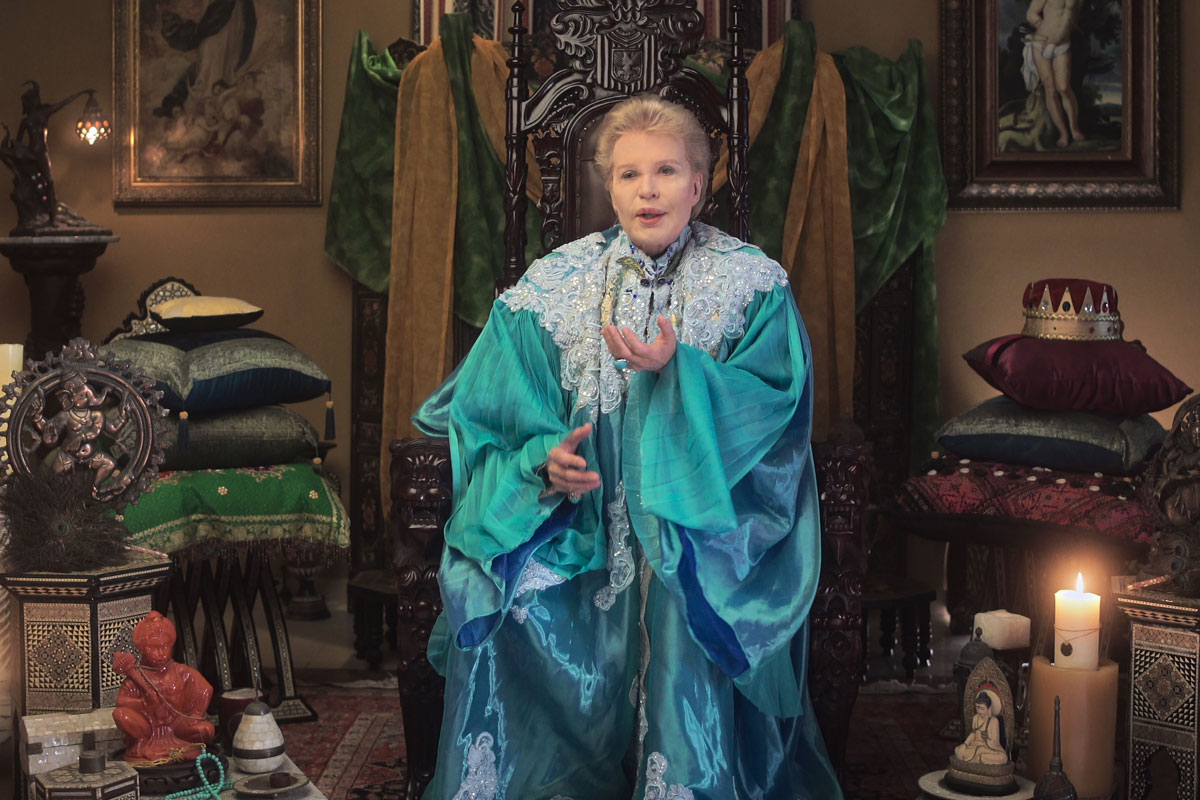 A light-haired sits on an ornate chair in a teal satiny garment surrounded by cushions, hanging fabric, occult objects and a crown.