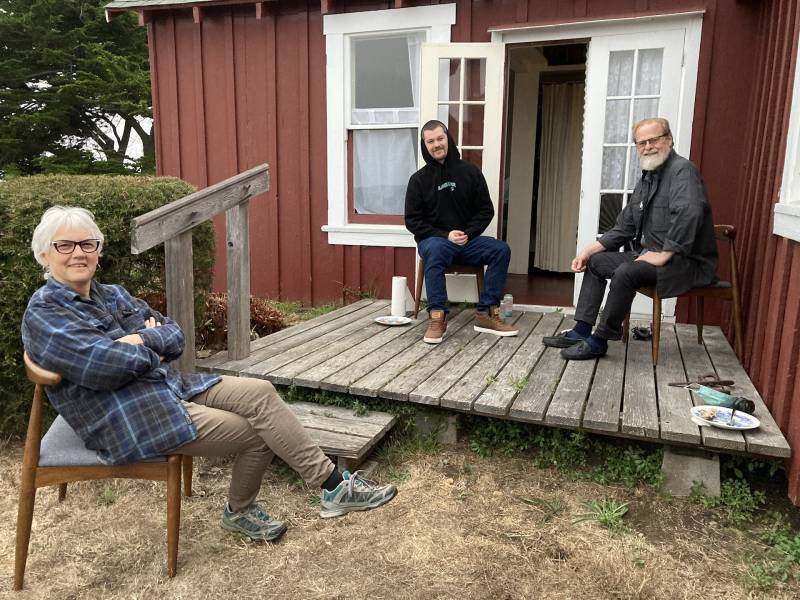 A family of three, two older adults and their adult son, sits outside a wooden house.
