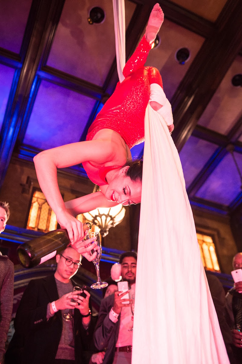 An aerialist hangs from silk while pouring champagne.