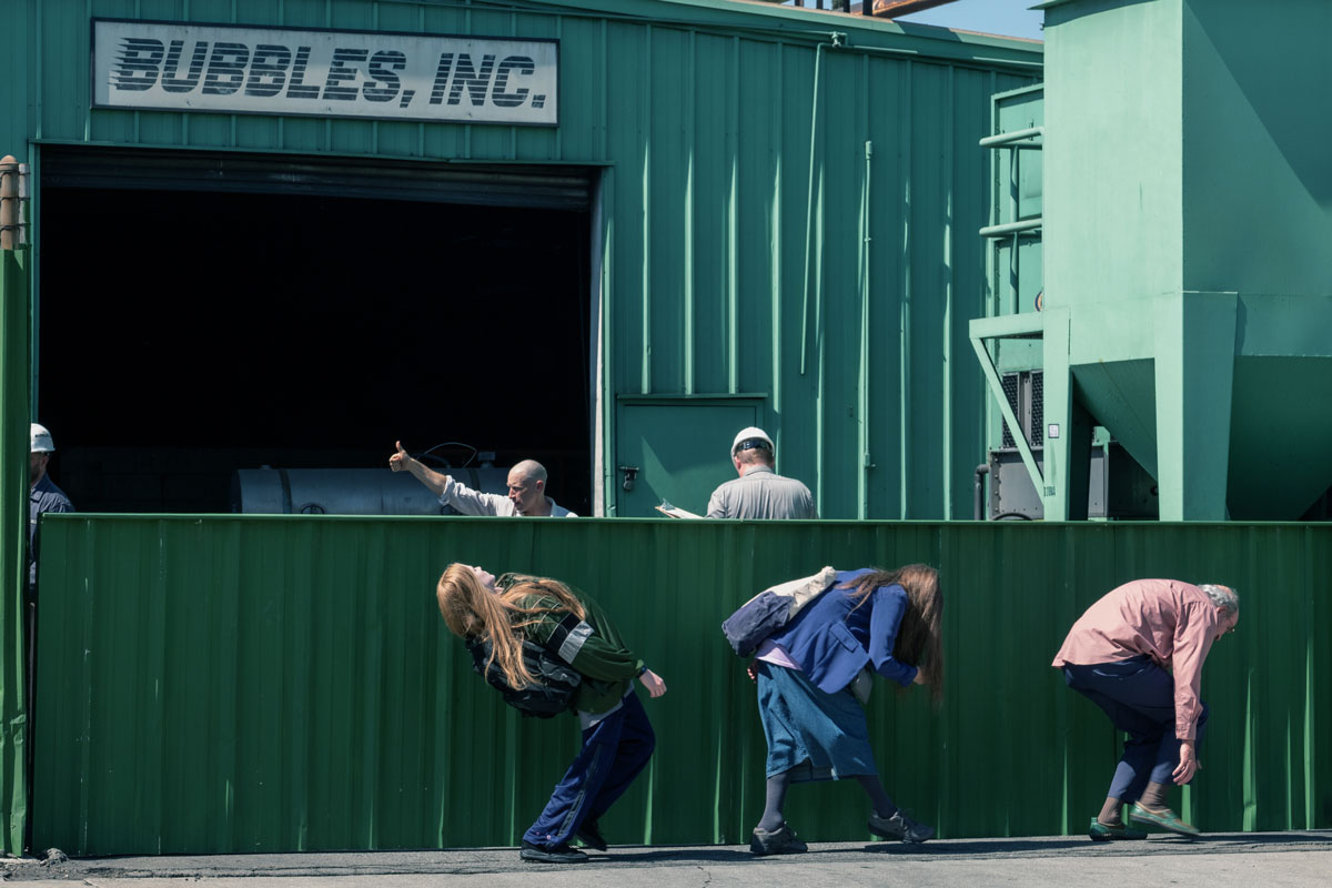 Three people bend to hide below a low wall outside a green building with a sign that says "Bubbles, Inc."