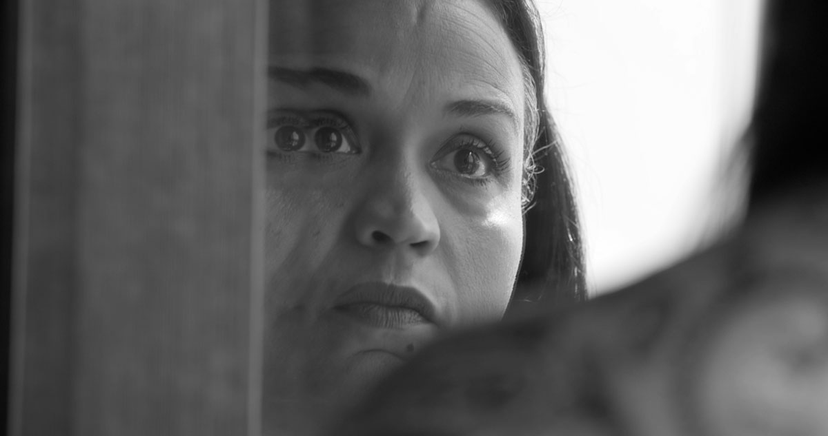 Black-and-white-image of a woman's face seen though a pane of glass over someone else's shoulder, distortion makes her left eye appear doubled.