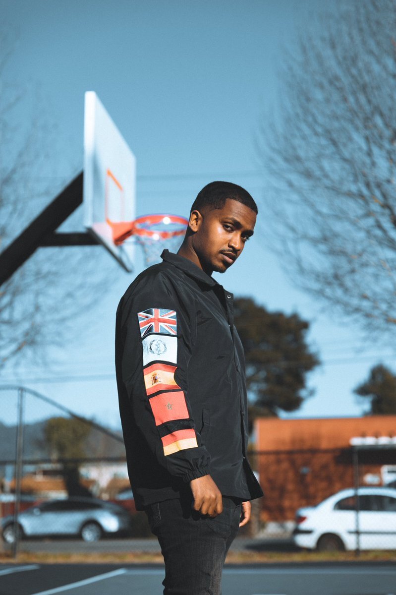 A Black man wearing a black bomber jacket with flags running down the arm stands in front of a basketball hoop. The camera looks up at him, and he looks down at the camera from a sideways glance. The trees are out of focus in the background, the bigger trees have already lost all their leaves and the sky is clear with no clouds. 