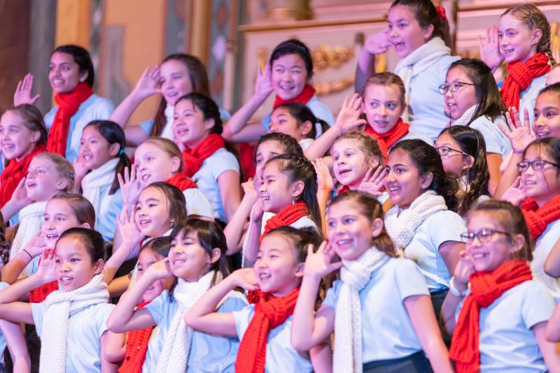 A chorus of young girls raise their hands to their ears while smiling.