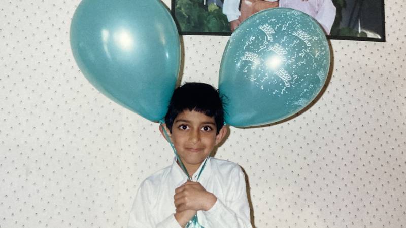 Young Indian boy looks straight into the camera while holding two light blue baloons that are larger than his head.