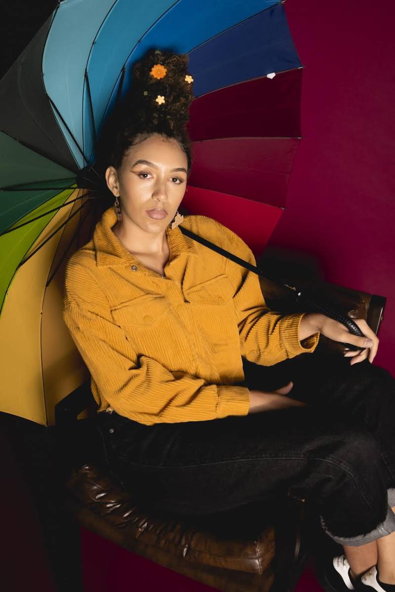 Jada Imani wears a mustard yellow jacket and poses holding a multicolored umbrella. 
