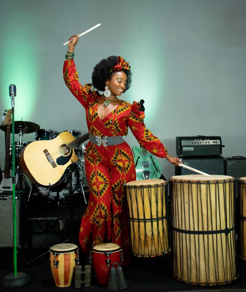 Queen Iminah plays the drums while wearing a guitar over her shoulder.