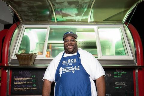 Champ Green stands in front of a food truck wearing a baseball cap and an apron.