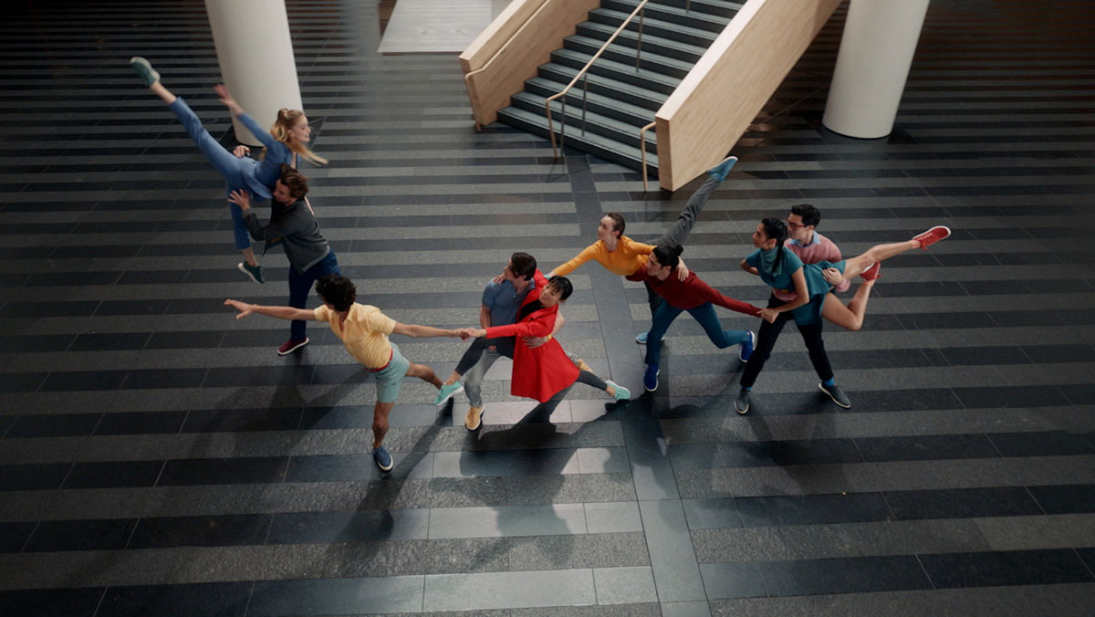 Colorfully dressed dancers holding hands extend across the gray and black-patterned floor of SFMOMA's lobby space.