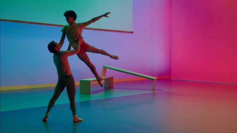 One male dancer holds another male dancer aloft in a teal and purple-tinged space.