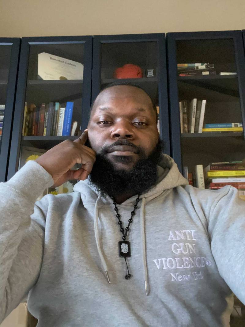 Antoine Johnson sits in front of a bookshelf, looking down at the camera, one hand supports his head thoughtfully, but he looks sharp and ready. His light gray sweatshirt says "Anti-Gun Violence". Behind him are shelves of books and certificates.