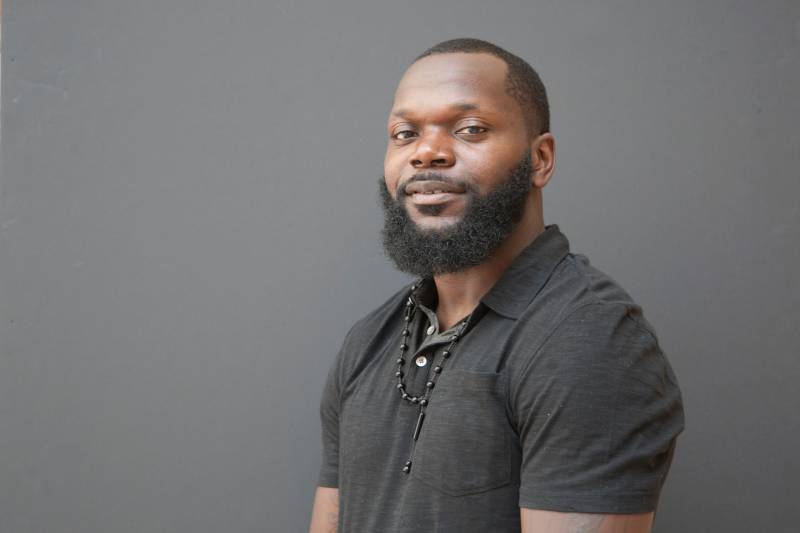 Antoine Johnson sits in front of a gray background wearing a dark gray polo T-shirt. His face is turned towards us with a slight smile and relaxed eyes. We can't see below his elbows but his arms seel to rest at his sides. His beard is neat and his hair looks freshly cut.