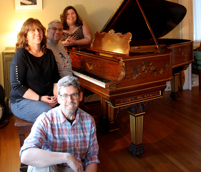 A family of two parents and two adult children pose around their grand piano in a cozy living room.