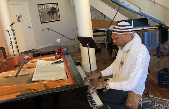 A man in a white sweater, jewelry and knit hat plays the piano. 