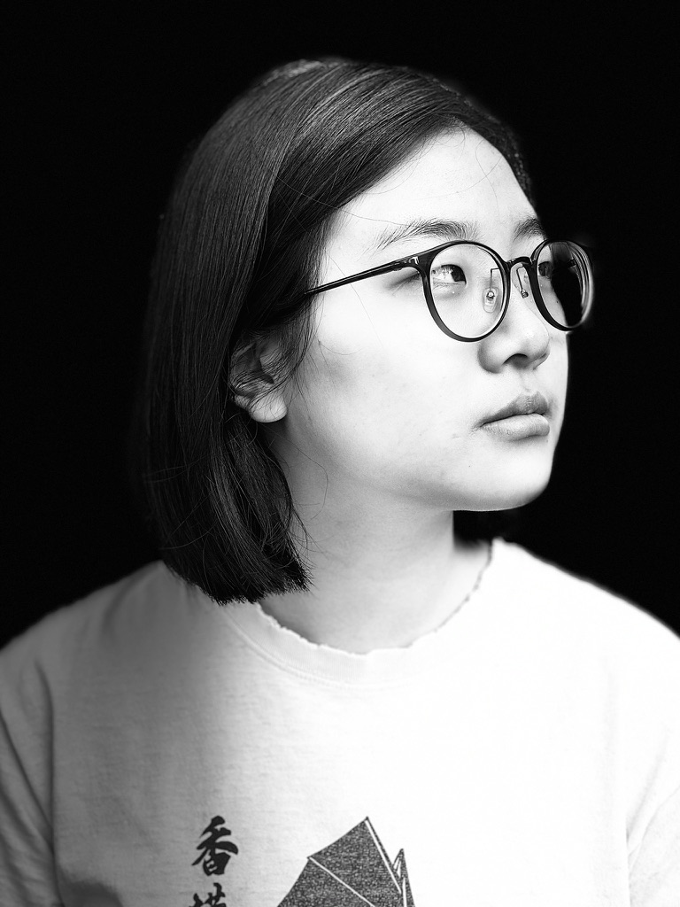 A young Asian American woman with glasses gazes into the distance in a back and white portrait.