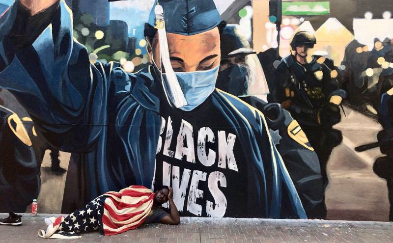 A man named Jeremiah sleeps wrapped in an American flag on the streets of downtown Oakland, beneath a large Black Lives Matter mural.