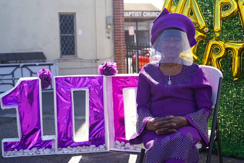 Marcella Hubbard, wearing all purple and a COVID-19 safe protective face shield, sits in front of a huge "100" sign as she celebrates her 100th birthday.