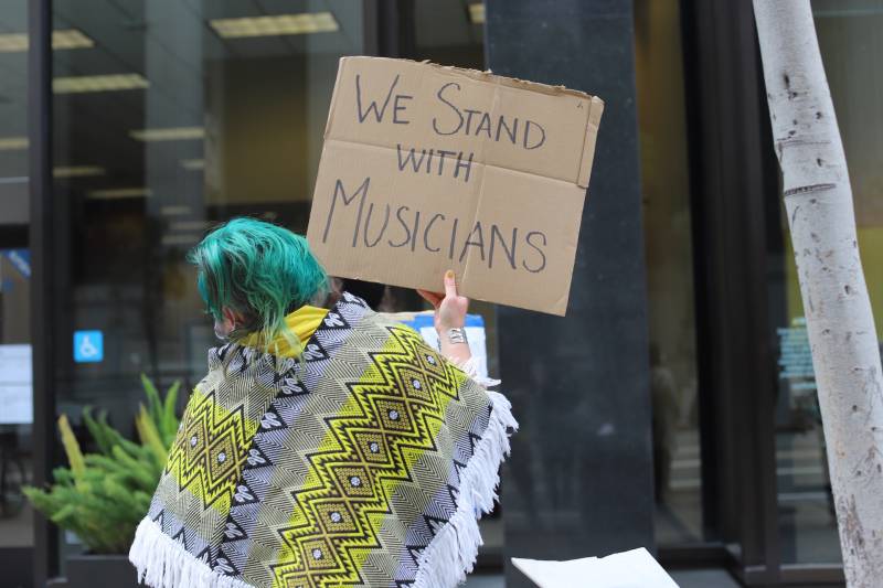 A woman with green hair holds a sign that says "We stand with musicians."