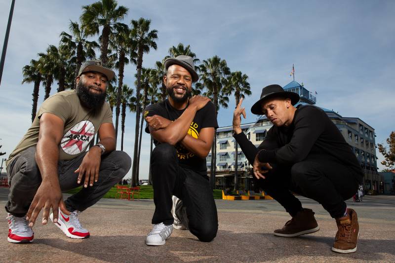 Three musicians smile and pose in front of palm trees.