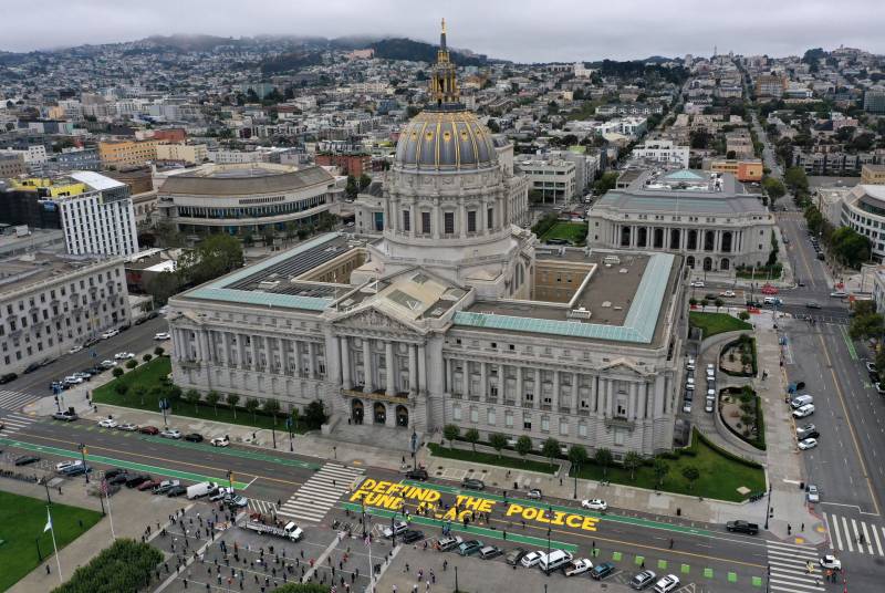 An aerial view of an ornate, official building with a large dome at its center. Yellow painted letters in the street in front of the building spell out "defund the police."