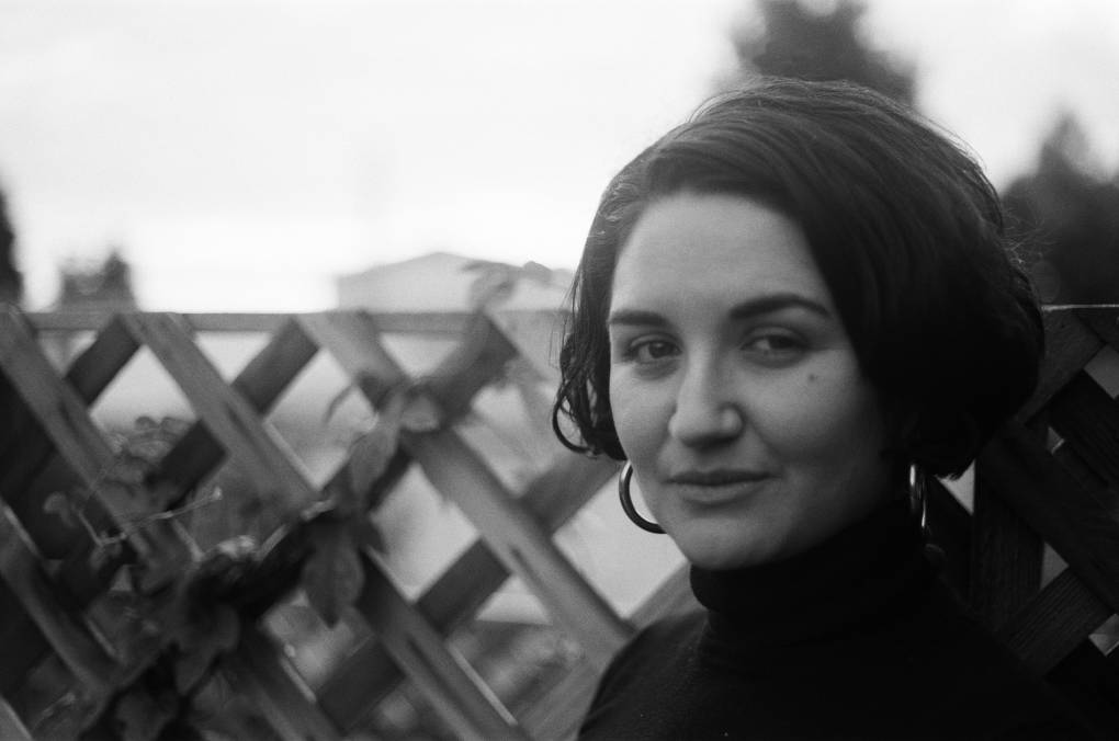 A black and white headshot of Maya Cueva. She gives us a half smile, is wearing hoop earings and her curly hair is in a bob. She wears a black turtleneck in front of a fence.