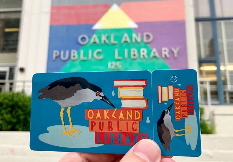 Someone holding a colorful Oakland Public Library card that has the image of a grey heron and a stack of books on it.