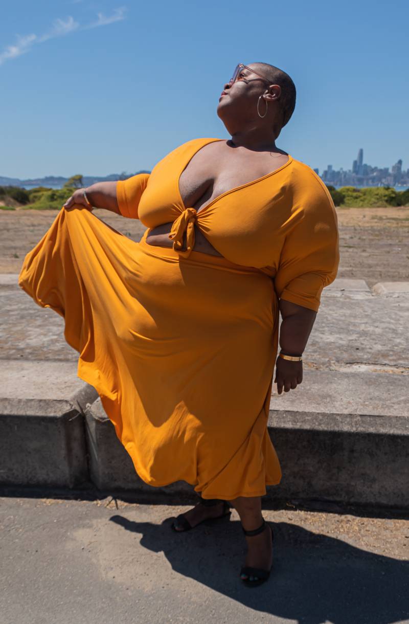 Brena Jean in matching yellow top and skirt. She stares up towards the sun, soaking up its rays while lifting a corner of her skirt.