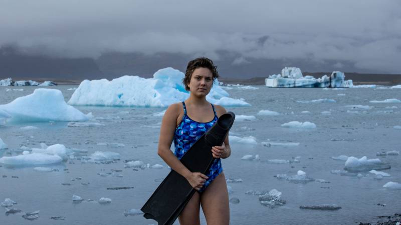 A young white woman with short brown hair wears a blue patterned one-piece bathing suit and holds large black flippers across her body. Behind her ice floats in water.