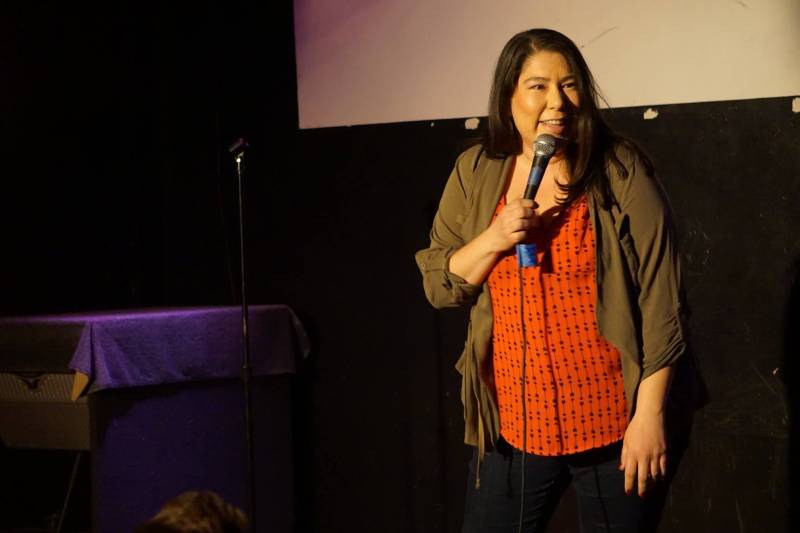Standup Comedian Jackie Keliiaa holds a microphone on stage, she leans forward slightly, smiling out at the audience.