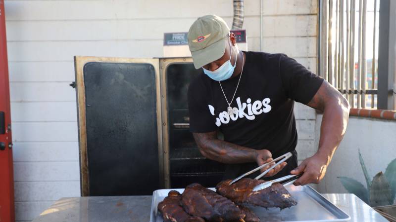Chef in a face mask and baseball cap lifting a rack of ribs with tongs.