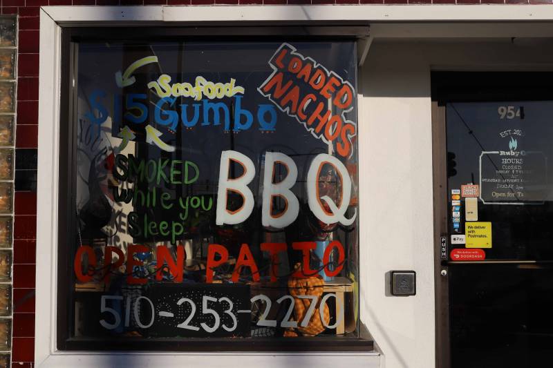 Writing in the restaurant window reads, "Seafood Gumbo," "Loaded Nachos," "Smoked While You Sleep," "BBQ," and "Open Patio."