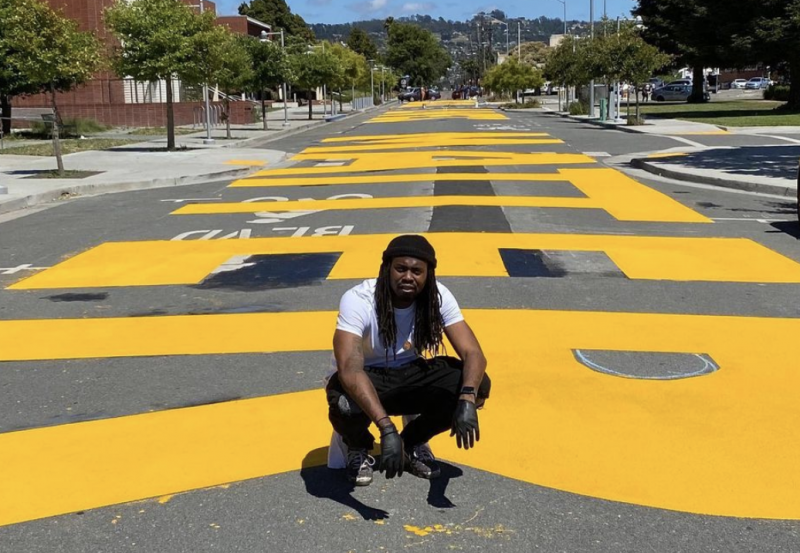 In June of 2020, Deonta Allen and community painted "Black Lives Matter" in bold bright yellow letters on the street in downtown Richmond, CA. Here he is, in a crouched position, posing in front of the finished painting.