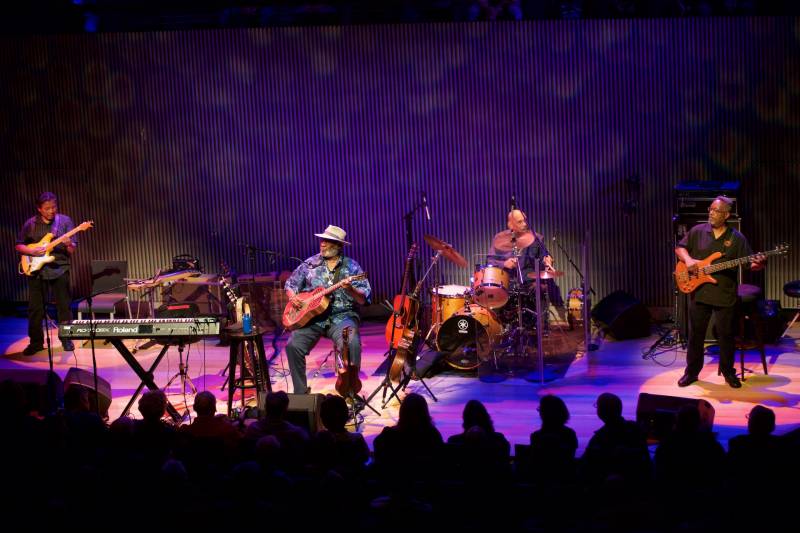 A jazz quartet performs onstage in a modern theater with purple lighting.