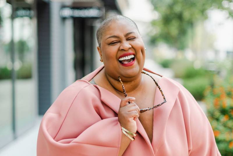 Brena Jean, advocate for those living with lipedema, takes a candid photo as she smiles and laughs while wearing a peach colored outfit and holding her glasses.