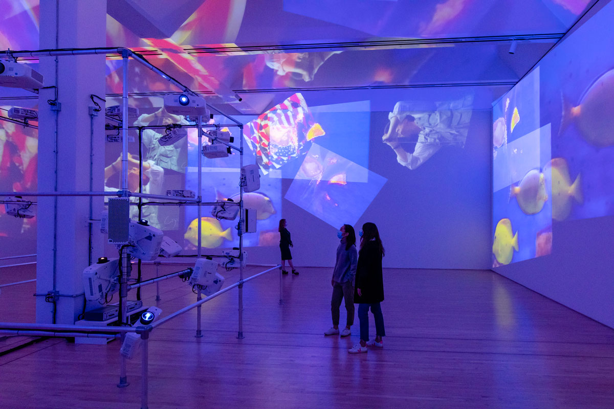In a large gallery filled with purple-blue light, multiple project images layer over the walls and ceiling as three people stand around watching.