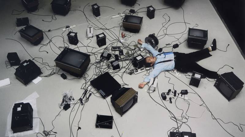 Seen from overhead, a man in light blue shirt and black pants lays spread-eagle surrounded by TVs and their cables