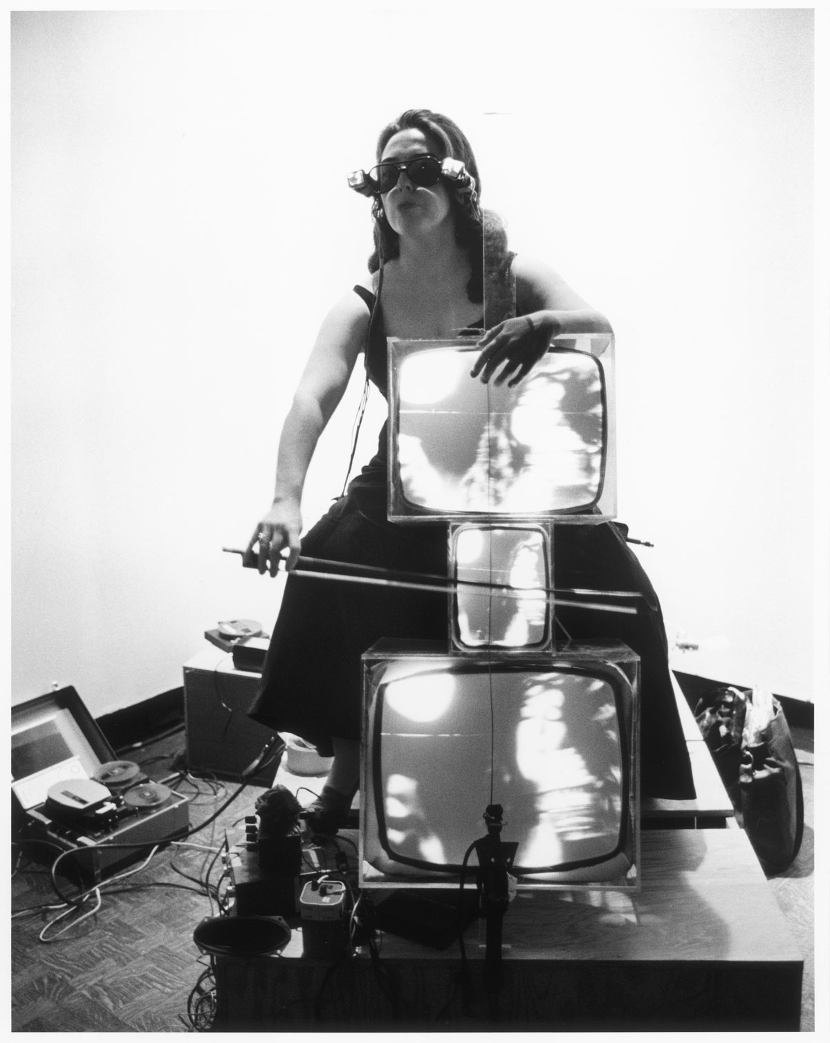 In a black-and-white photo, a woman wearing glasses made from small TVs plays a "cello" made from three TV monitors.