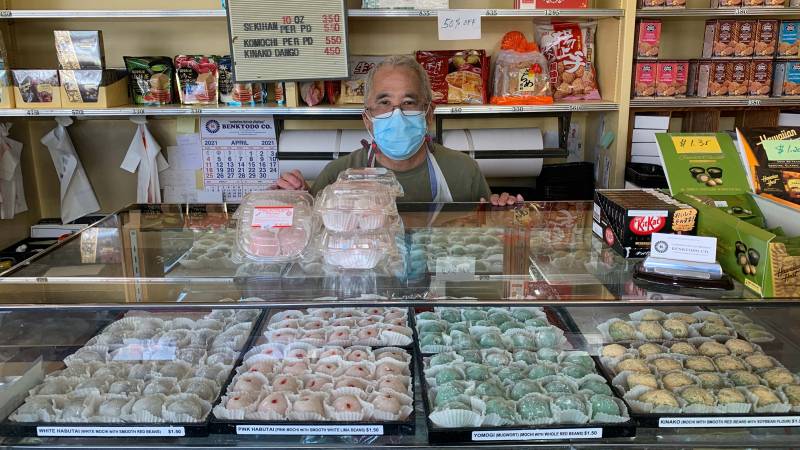Mochi shop owner stands behind display case while wearing a face mask.