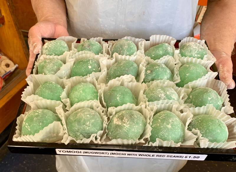 A man holds a tray of green mochi in paper sleeves.