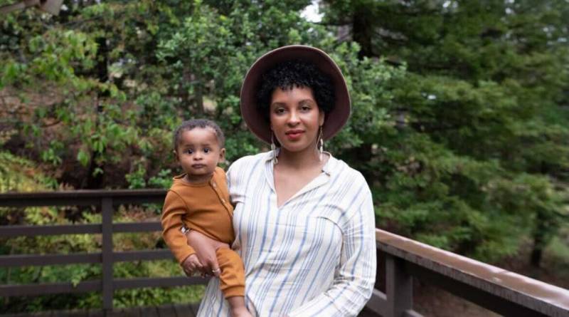 Brittany Tanner wears a white and grey striped shirt and a brown hat as she stands holding her son, Akai.