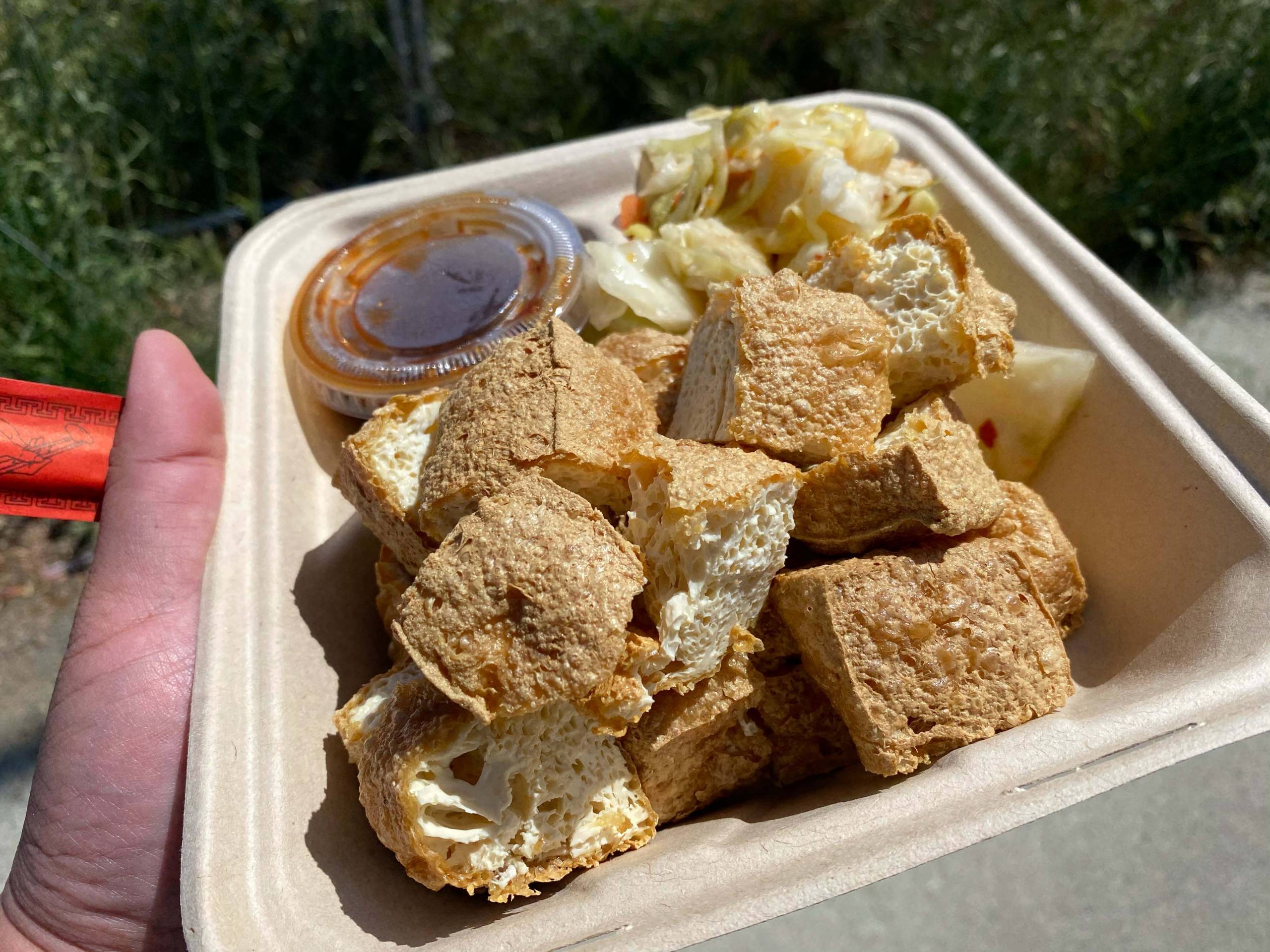 A takeout container of fried stinky tofu, with pickled cabbage and a tub of sauce on the side.