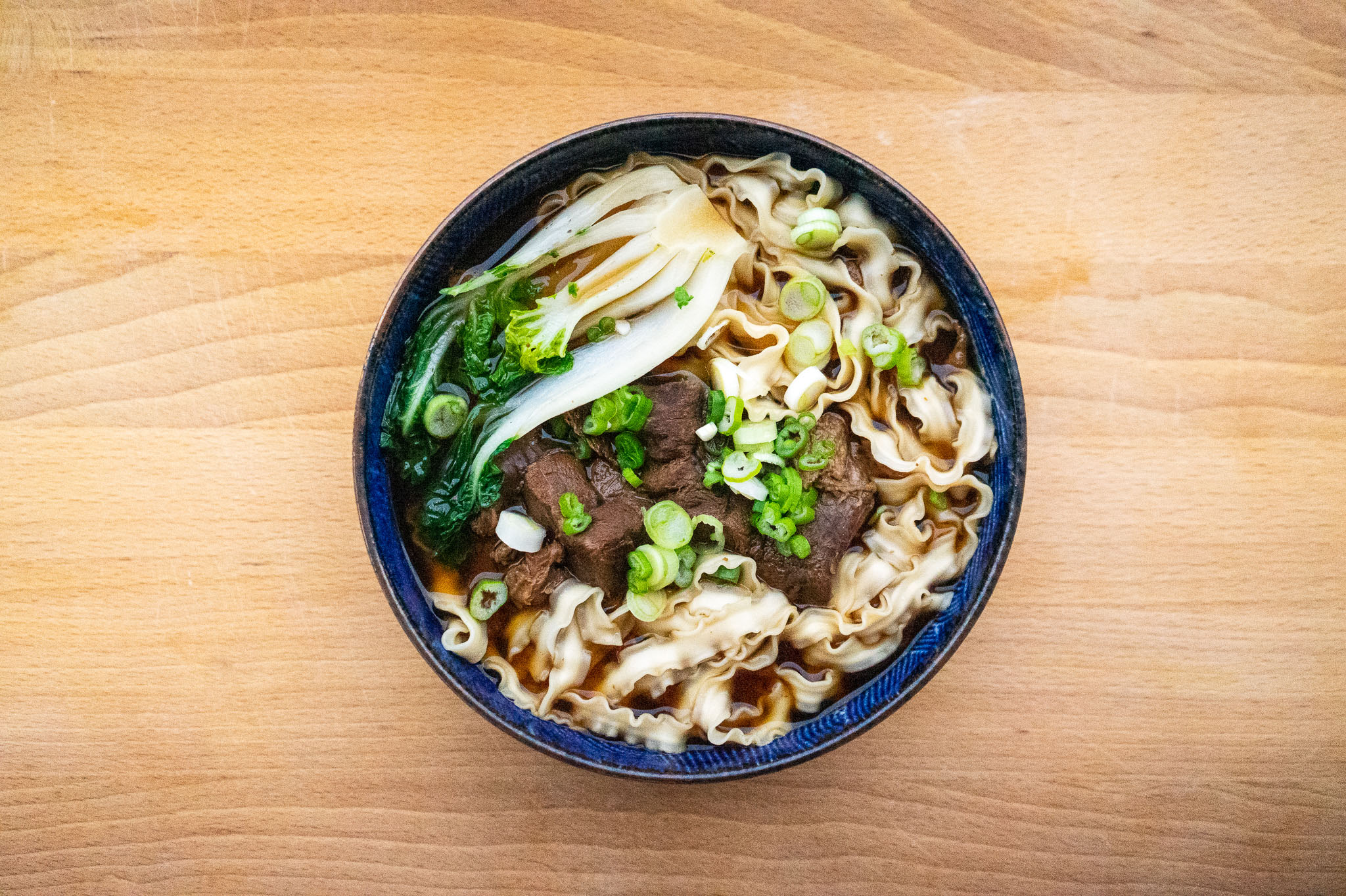 Overhead view of a bowl of beef noodle soup, garnished with bok choy and chopped scallions, on a wood table.