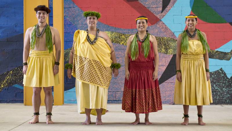 Four hula dancers in traditional attire pose in front of a colorful mural.
