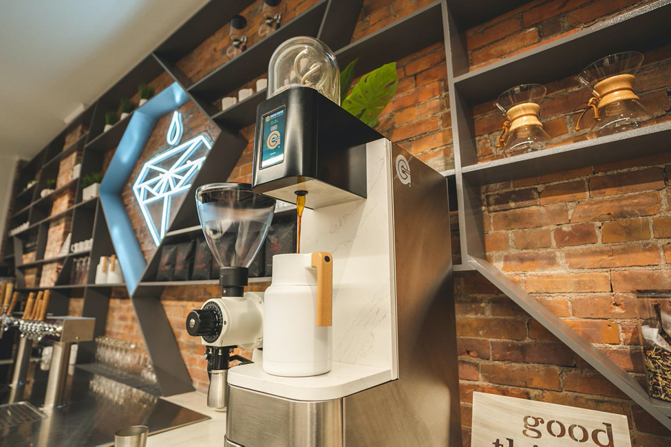 A Ground Control Coffee batch brewer in a coffee shop in front of an exposed brick wall with shelving.