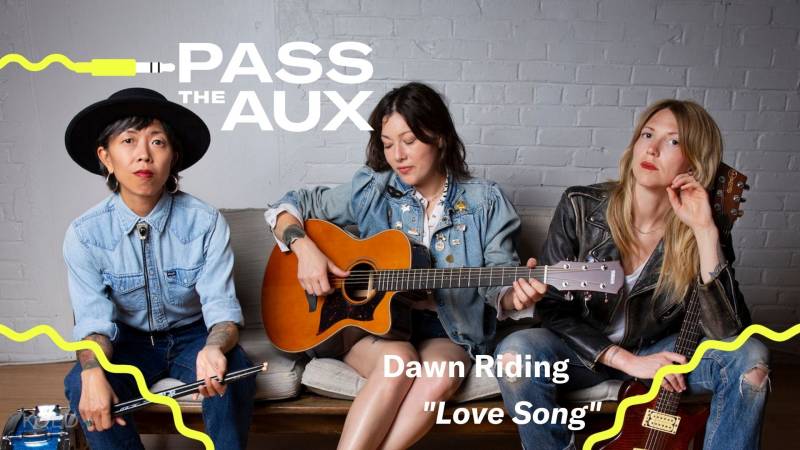 A musical trio dressed in denim sits on the couch; the woman on the left holds a drum stick, the one in the center strums an acoustic guitar and the one on the right holds an electric guitar.