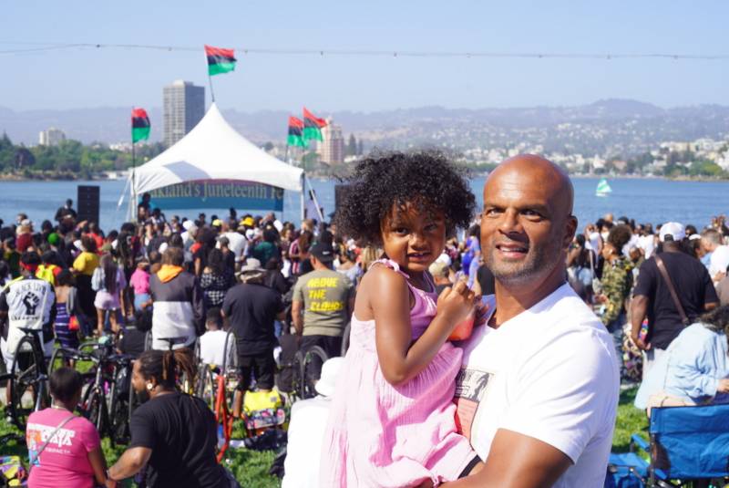 Travis Watts, organizer of Oakland's official Juneteenth celebration at Lake Merritt's amphitheater, is pictured with festival goers in the background as he holds his daughter Zaya.