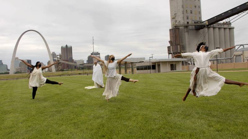 Three woman dancers on a grassy field, wearing matching off-white blouses and flowing white skirts, are in synchronized flying pose, against a backdrop of the St. Louis skyline and iconic St. Louis Gateway Arch.