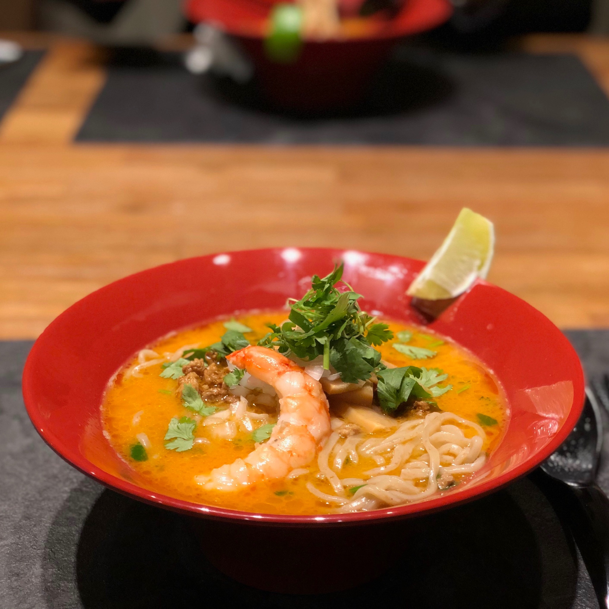 A bowl of Thai-inspired tom yum paitan ramen in a red bowl, topped with a large shrimp and a lime wedge.