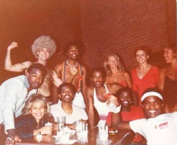 The cast of Dancing Wheels in 1979 at the end of a table, eleven of them stare into the camera. They are peak late seventies style, with big smiles.