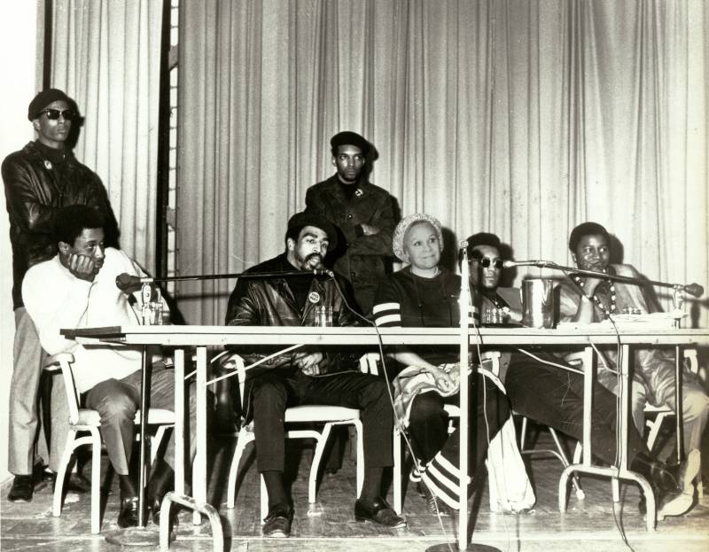 Katherine Dunham sits at a press conference table beside Reverend Charles Koen, Poet Eugene Redmond, and two members of the Black Eqyptians street gang.